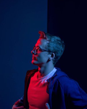 Siyah ve mavi arka planda duran ve bakan gözlüklü beyaz bir genç.