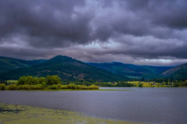 COUER D\'ALENE RIVER WITH A TREE LINED SHORE, MOUNTAINS AND A CLOUDY SKY