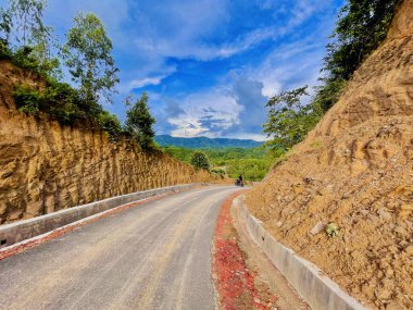 İki sarp tepenin arasında kıvrımlı bir yol. Etrafı yemyeşil, bulutlarla dolu parlak mavi bir gökyüzünün altında.