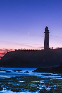 Bir deniz fenerinin dikey görüntüsü. Mor gökyüzü ve yarım ay.