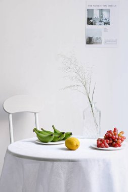 Dikey bir sandalye ve meyve dolu bir masa ve beyaz bir duvarda cam bir vazo.