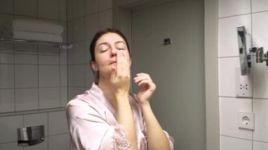 Genç bir dişi, gece kendi kendine bakım rutinini yapıyor ve yüzüne banyoda masaj yapıyor.