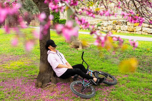 一个年轻人骑着自行车躺在一棵开着粉红色花的树下休息 春天的绿色城市概念 — 图库照片