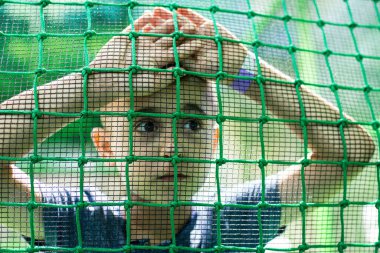 Küçük bir çocuk tek başına duruyor ve yeşil çitlere yaslanıyor.