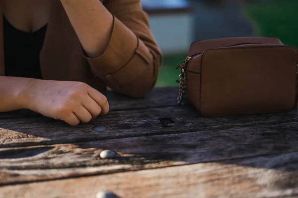A brown small bag on a wooden old table and a female next to it wearing a brown jacket