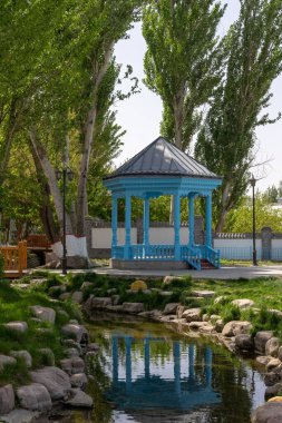 Parktaki mavi bir çardağın dikey görüntüsü küçük bir gölete yansıyor.