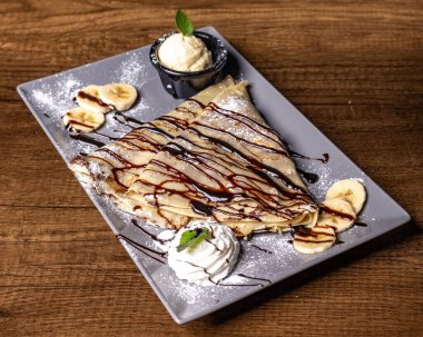 Çikolata soslu bir tabak krep, ahşap bir masada krem şantili muz dilimleri.