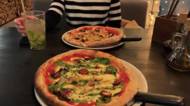 Masada iki pizza ve arkada çizgili tişörtlü bir kadın.