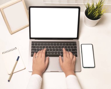 Dizüstü bilgisayar ve cep telefonu kullanan ve ofiste boş ekranı olan bir kadın elinin üst görüntüsü.