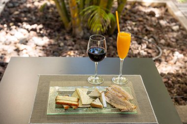 Üzerinde farklı peynir çeşitleri olan cam bir tabak ve masada bir bardak şarap ve meyve suyu.