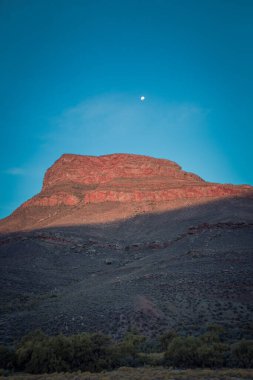 Akşam gökyüzünde ay olan bir dağ manzarasının dikey görüntüsü.