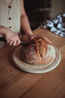 Yuvarlak bir tahtanın üzerinde taze pişmiş ekmek dilimlemek için bıçak kullanan bir kadın.