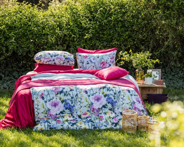 A bright shot of a bed with pink and floral bedsheets and pillows on the grass in a yard