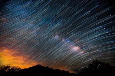 Gün batımında yıldızlarla dolu karanlık bir gökyüzünün altında dağ ve ağaçlardan oluşan bir siluet.