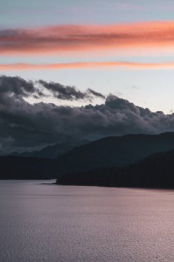 Estetik bulutlu, turuncu bir gün batımının dikey çekimi dağlar ve bir göl üzerinde.