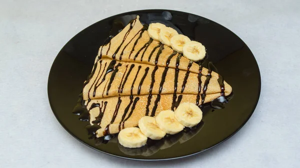 A closeup shot of crepes with chocolate syrup and bananas on a black plate