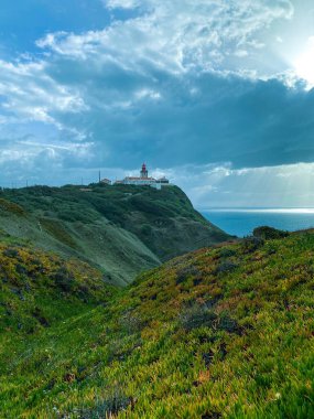 Güneşli, bulutlu bir deniz kıyısında deniz feneri olan tepelerin manzarası.