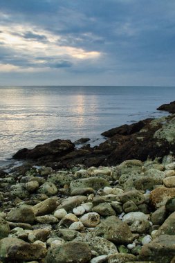 Gün batımında bulutlu bir gökyüzünün altında kayalık bir sahilin dikey çekimi.