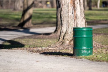 Parktaki ağacın dibinde duran yeşil metal bir çöp tenekesi.