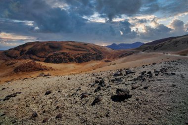 Teide Dağı 'ndaki çorak bir volkanik manzara dramatik bulutlu bir gökyüzünün altında kayalık, kırmızımsı kahverengi tepeleri gösteriyor..