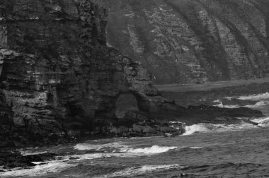 Kıyı şeridinin gri tonlu görüntüsü kayalık dağlara çarpıyor.