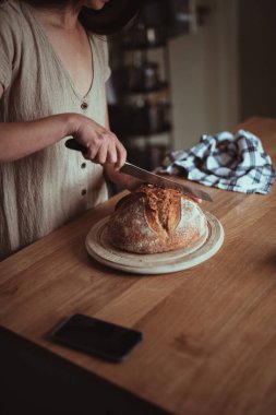 Yuvarlak bir tahtanın üzerinde taze pişmiş ekmek dilimlemek için bıçak kullanan bir kadın.