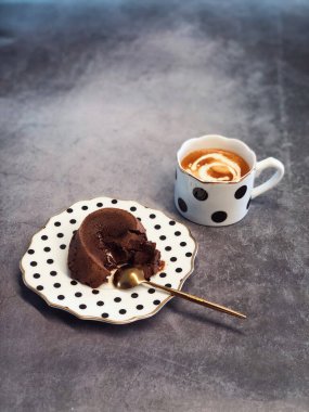 Bir fincan sütlü kahveyle noktalı tabakta duran çikolatalı lava kekinin dikey görüntüsü.