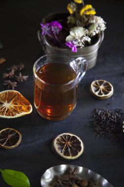 Koyu ahşap bir yüzey üzerinde kuru limon ve bitkilerle dolu bir fincan çayın dikey yakın çekimi.
