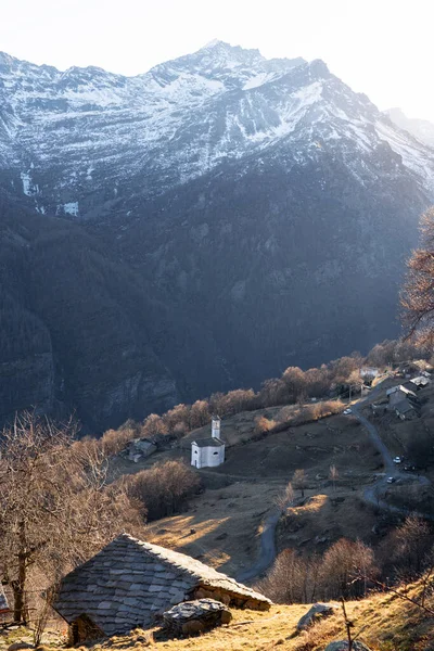 A vertical shot of snow-covered mountains with a few buildings on the slopes on a sunny day