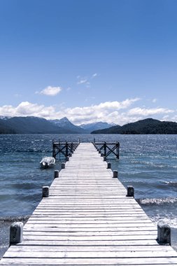 Arka planda dağlar olan göldeki ahşap bir iskelenin dikey görüntüsü.
