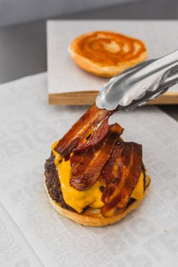 Lezzetli bir hamburgerin üzerine domuz pastırması koyan dikey bir maşa.