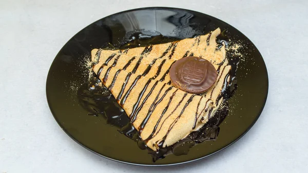 A closeup shot of crepes with chocolate syrup and cookie on a black plate