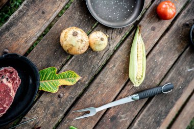 Çiğ biftek, soğan, mısır ve barbekü çatalıyla ahşap piknik masasının üst görüntüsü.