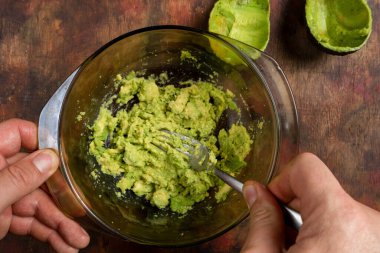 Tahta bir masada izole edilmiş küçük bir kasede avokadoyu ezen ellerin üst görüntüsü.