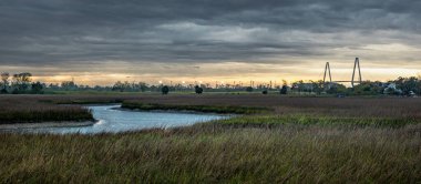 Gün batımında bulutlu bir gökyüzünün altında akan bir nehrin manzarası.