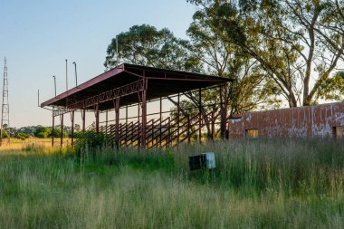 Güneşli bir günde yeşil çimen ve ağaçlardan oluşan bir alanda metalik bir gölgeliğin manzarası.