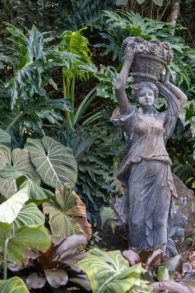 A vertical shot of a metal statue of a woman carrying a basket on her head surrounded by lush greenery
