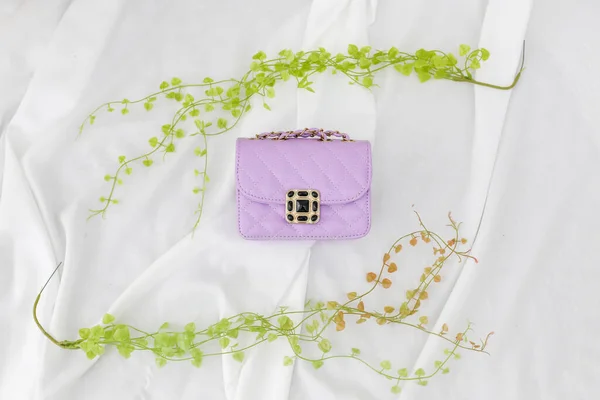 植物で飾られた柔らかい白い生地に紫の女性の革のバッグのトップビュー — ストック写真