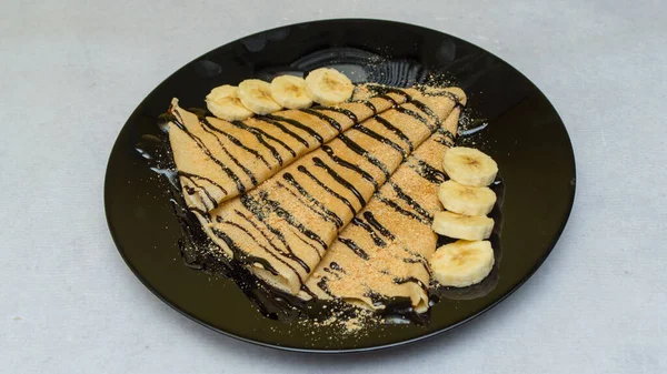 A closeup shot of crepes with chocolate syrup and bananas on a black plate