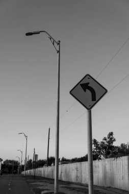 Bir sokak lambasının yanındaki direkte trafik işaretinin ilerisindeki sol virajın gri tonlu görüntüsü.