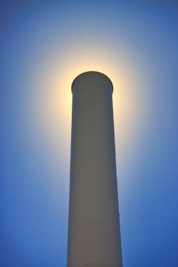 Açık mavi gökyüzünün altında güneşi kaplayan fabrika bacasının dikey minimalistik görüntüsü.