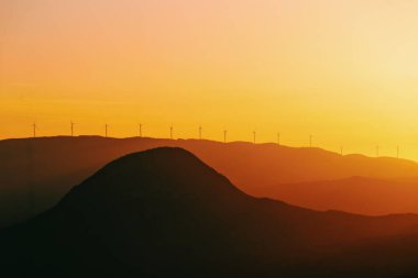 Gün batımında dağlarda sisli bir havanın silüeti ve tepesinde yel değirmenleri.