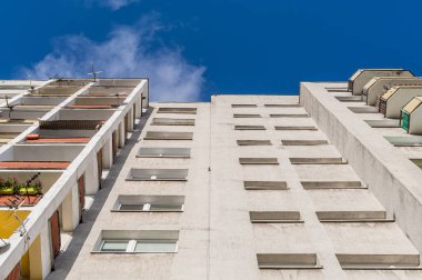 Bulutlu bir gökyüzüne karşı pencereleri ve balkonları olan yüksek bir binanın dış yüzeyi.