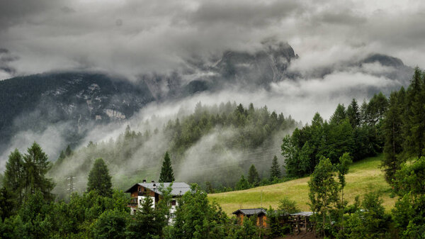 A scenic shot of village wooden huts surrounded by forests on a mountain under the clouds