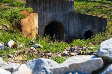 Etrafı kayalar ve yeşil bitkilerle çevrili bir kanalizasyon tüneli.