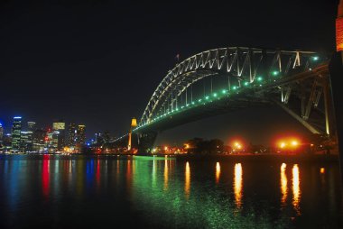 Sydney Limanı Köprüsü 'nün düşük açılı bir görüntüsü. Işıklar geceleyin nehire yansıyor.