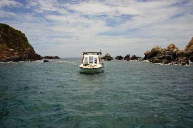 Bulutlu gökyüzünün altındaki kayaların arasında denizde beyaz bir tekne görüntüsü.