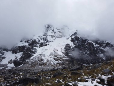 Bulutlu bir gökyüzünün altında karla kaplı kayalık bir dağın manzarası.