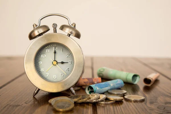 A closeup shot of a clock with money aroung on a wooden table - \
