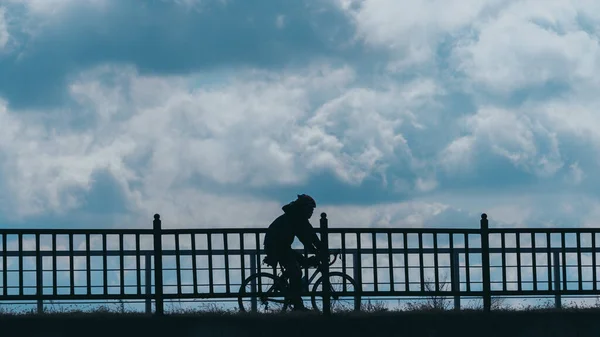 A silhouette of a male riding a bicycle on a bridge with the cloudy sky in the background